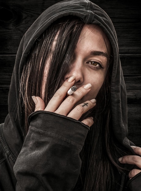 Portrait of a young girl with long hair in a hood that holds in her fingers and smokes a cigarette