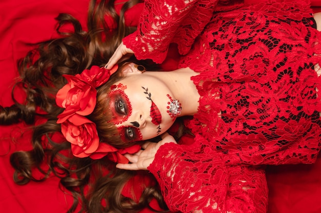 Portrait of a young girl with make-up and Dia de los Muertos attire lying on a red background.