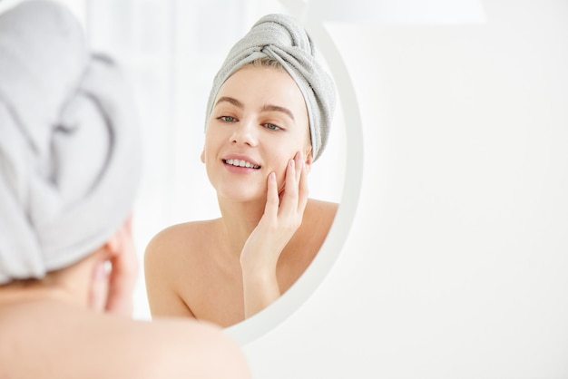 Portrait of young girl with towel on head in white bathroom looks and touches her face in the mirror and enjoys youth and hydration Natural beauty home care for problem skin