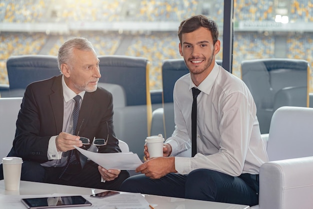 Portrait of young guy and his mature coworker sitting at table in stadium office discussing business documents and drinking coffee