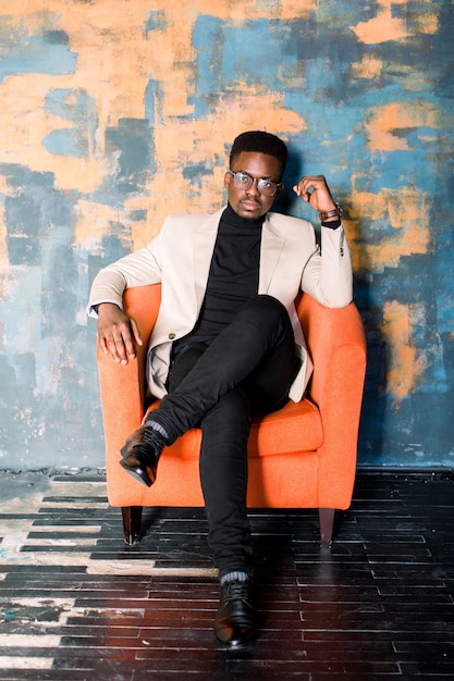 Portrait of a young handsome african american man in an elegant suit and glasses sitting in an orange armchair on a grunge background. Studio shot.