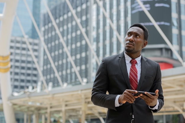 Portrait of young handsome African businessman in suit against view of modern building in the city outdoors