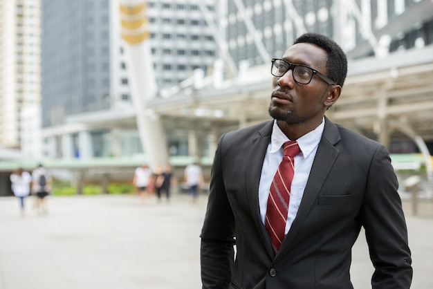 Portrait of young handsome African businessman in suit against view of modern building in the city outdoors