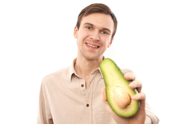 Portrait of young handsome caucasian brunette man smiling and showing half an avocado isolated on white studio background