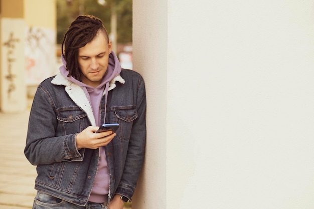 Portrait of young handsome man with dreadlocks browsing mobile phone standing leaning against wall on street