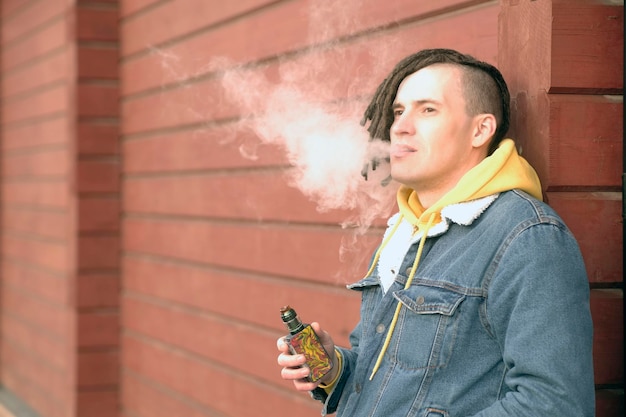 Portrait of young handsome man with dreadlocks vaping standing leaning on wall on city street Adult guy smoking ecigarette letting off steam