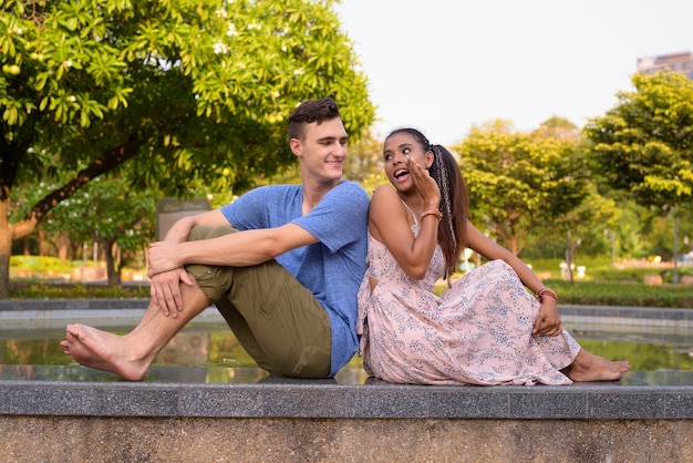 Portrait of young handsome man and young beautiful Asian woman relaxing together at the park