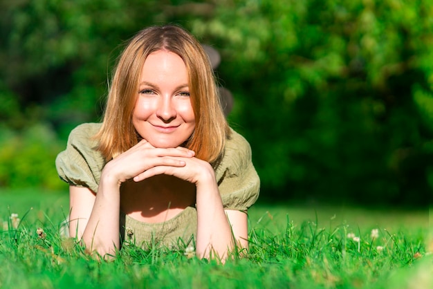 Portrait of young happy cheerful positive teenager girl, young pretty joyful woman is lying on stomach on green grass, smiling, looking at camera, enjoying summer good day. Copy space, place for text