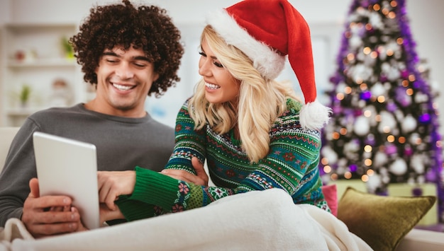 Portrait of young happy couple celebrating Christmas at home, relaxing and looking at something fun on tablet.