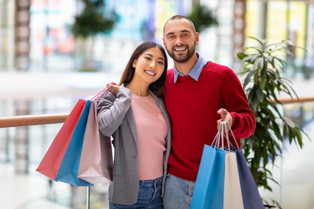 Portrait of young happy diverse couple posing with shopper bags smiling at camera at huge city mall