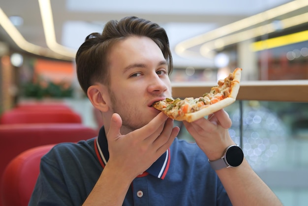 Portrait of young happy handsome man hungry guy is sitting on food court in shopping mall eating holding in hands juicy tasty piece of pizza biting Fast junk unhealthy fat food Hunger