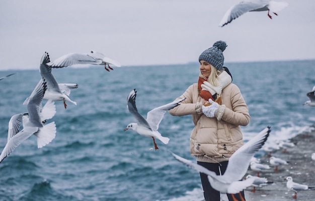 Portrait of young happy woman feeds seagulls on the sea Pretty female wearing coat scarf hat watching flying seagulls by the sea on cold season People and nature concept