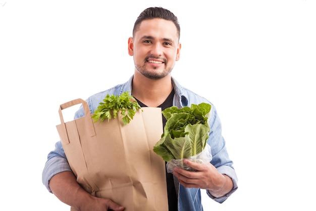 Portrait of a young Hispanic man carrying a bag of groceries with a lot of green in it against a white background