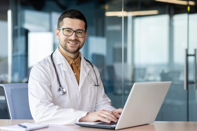 Portrait of a young intern specialist doctor sitting in the office at the table studying working at the laptop in a white coat and with a stethoscope and looking at the camera with a smile