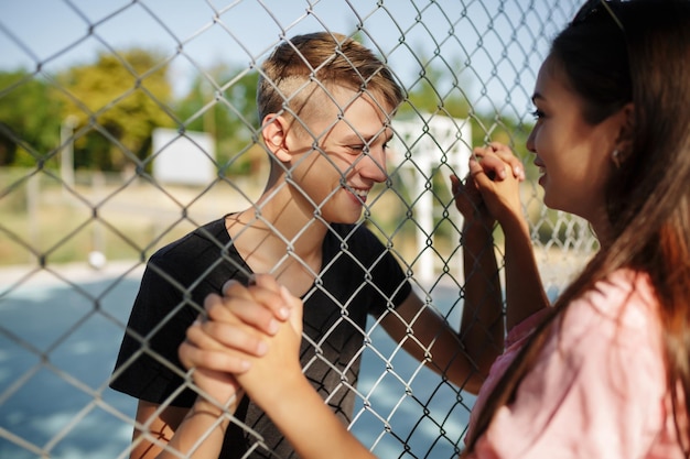 Portrait of young joyful boy standing on basketball court while happily looking at nice girl with dark hair through mesh fence and holding her hand