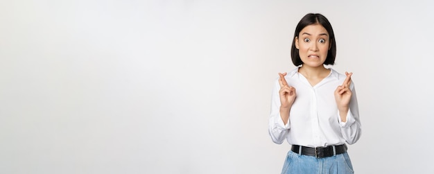 Portrait of young korean woman asian girl cross fingers and praying making wish anticipating waiting for results standing over white background