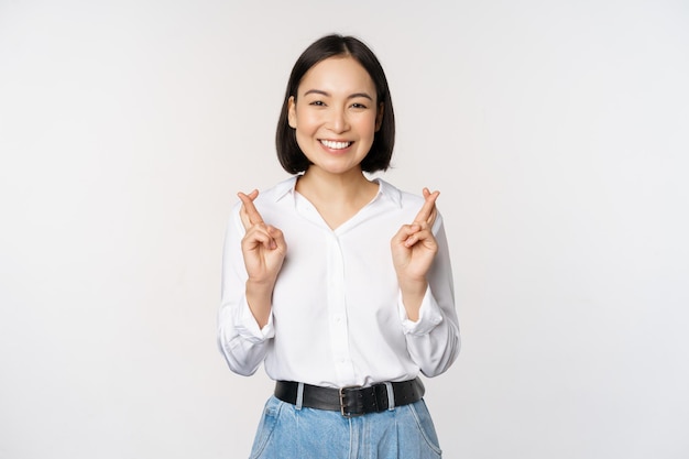 Portrait of young korean woman asian girl cross fingers and praying making wish anticipating waiting for results standing over white background