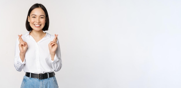 Portrait of young korean woman asian girl cross fingers and praying making wish anticipating waiting for results standing over white background