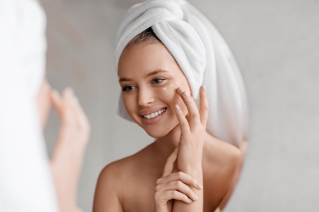 Portrait of young lady applying nourishing eye cream after shower looking at mirror and smiling in