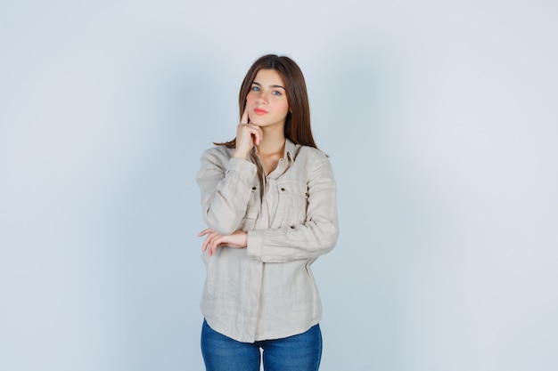 Portrait of young lady keeping finger on cheek in casual, jeans and looking confident front view