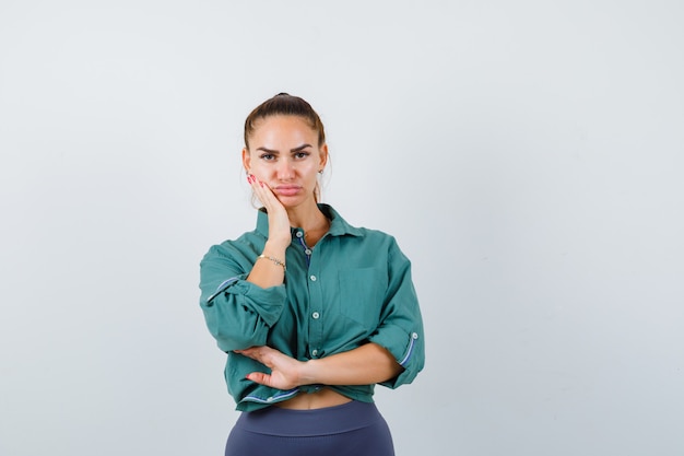 Portrait of young lady leaning cheek on hand in green shirt and looking gloomy front view