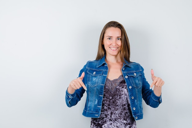 Portrait of young lady pointing down in blouse, denim jacket and looking joyful front view