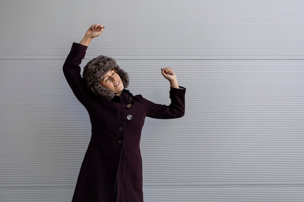 Portrait of a young Latin American woman 22 standing with outstretched arms with positive expression wearing a purple coat with imitation fur hat on a gray background