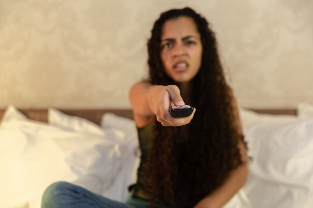 Portrait of young latin woman with remote lying in bed and watching TV. 