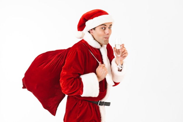 Portrait of young man 30s in santa claus costume and red hat holding gift bag and drinking champagne