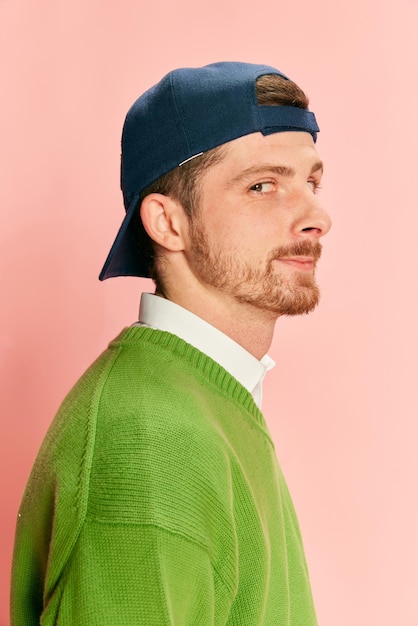 Portrait of young man in cap posing in green sweater and shirt isolated over pink background Side view