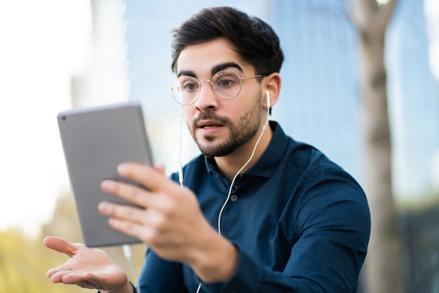 Portrait of young man having a video call on digital tablet while standing on bench outdoors. Urban concept.