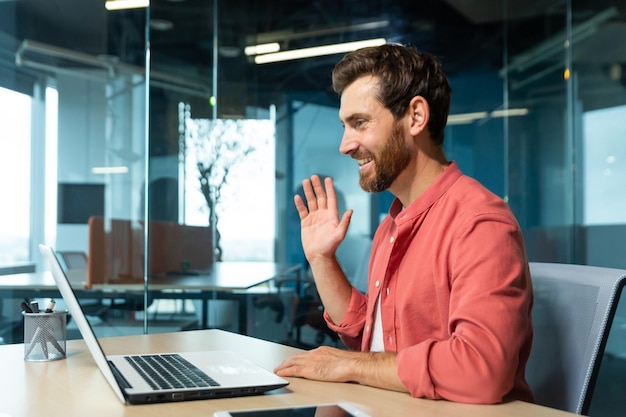 Portrait of a young man in a red shirt sitting in the office at the table working on a laptop