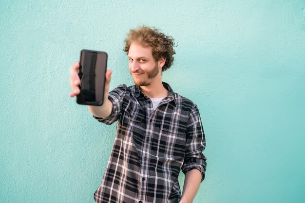 Portrait of young man showing blank smartphone screen against light blue wall.