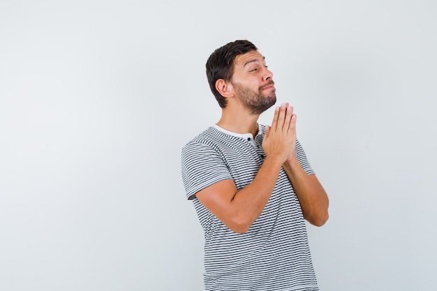 Portrait of young man showing clasped hands in pleading gesture in t-shirt and looking hopeful front view