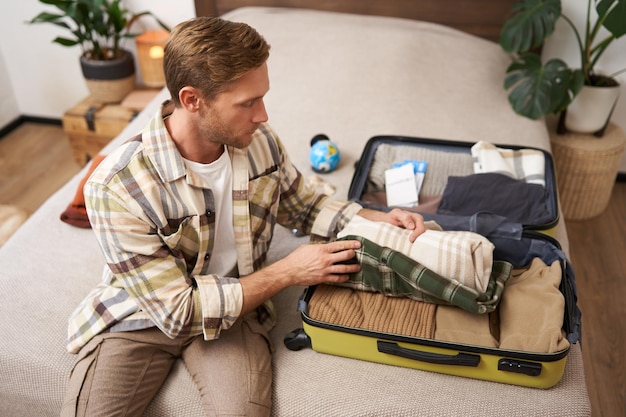 Photo portrait of young man sits on bed with suitcase goes on holiday packs clothes for vacation or