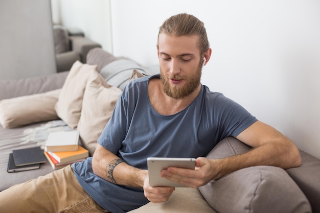 Portrait of young man sitting on gray sofa with earphones and the tablet in hands at home