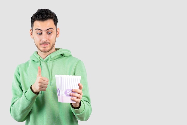 Portrait of young man in sweatshirt holding popcorn box and giving thumbs up