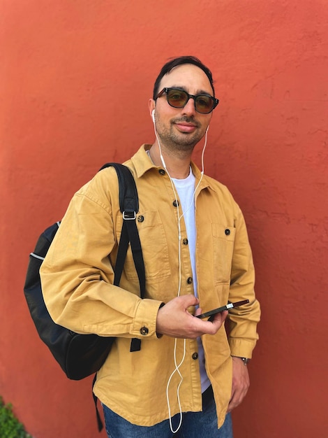 Portrait of a young man who stands on the background of a orange wall listening to music in wired headphones