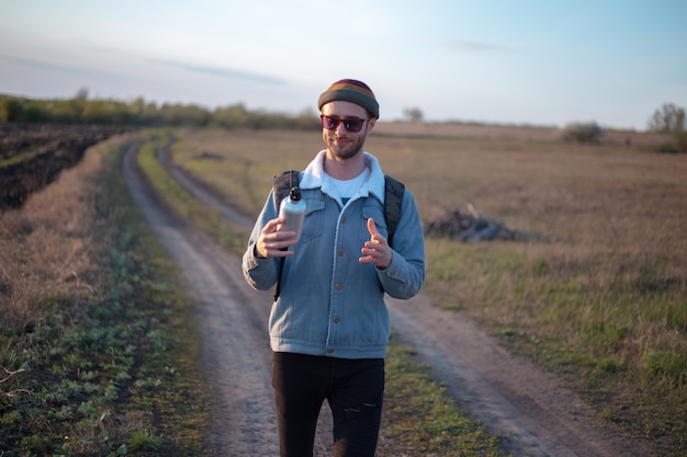 Portrait of young man with backpack holding reusable aluminum thermo water bottle in hand