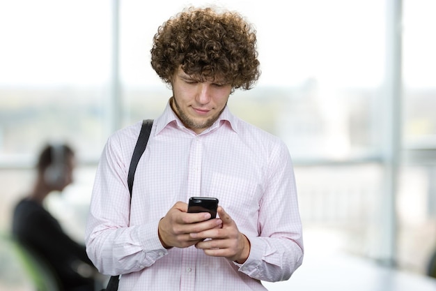 Portrait of a young man with curly hair using his smartphone in the office indoor window in the