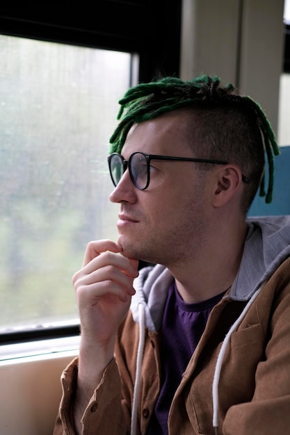 Portrait of young man with green dreadlocks in glasses riding on train looking at nature Close up