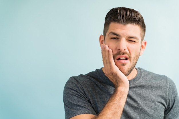 Portrait of young man with hand on cheek suffering from toothache against turquoise background