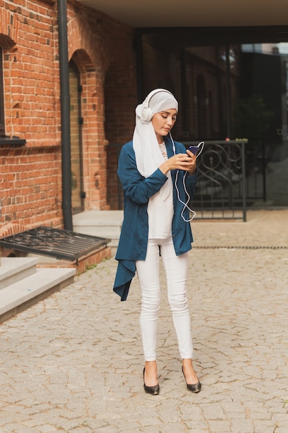 Portrait of young modern arabian woman holding mobile phone and listening the music to headphone