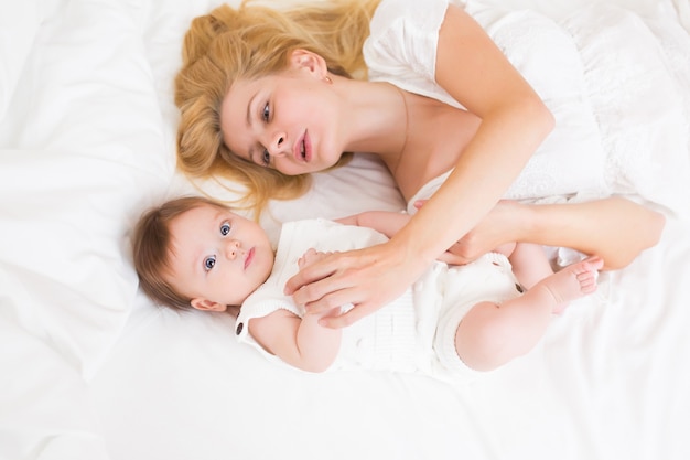 Portrait of young mother with blond hair with her sweet 3 month old baby girl in white wear having fun in the bedroom at morning, loving happy family concept, top view