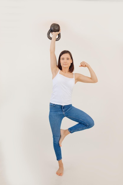 Portrait of young muscular woman wearing white top blue jeans standing on leg lifting kettle bell sh