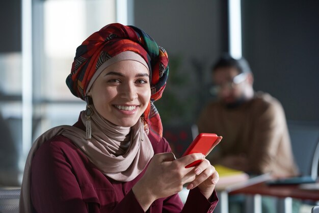 Portrait of young muslim businesswoman smiling at camera while reading a message on her mobile phone at office