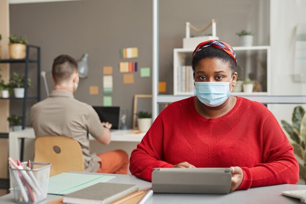 Portrait of young overweight woman in protective mask looking at front while sitting at workplace and working on tablet pc at office