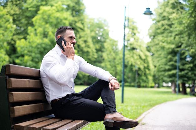 Portrait Of A Young Physically Fit Businessman Smokes Cuban Cigar in Park and Talks on Mobile Phone