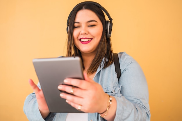 Portrait of young plus size woman listening to music with headphones and digital tablet outdoors on yellow