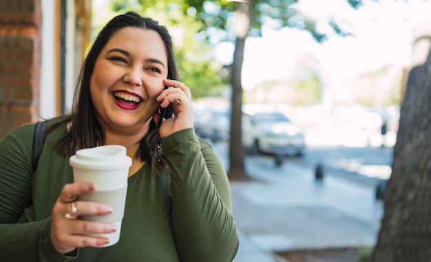 Portrait of young plus size woman talking on the phone while holding a cup of coffee outdoors in the street.